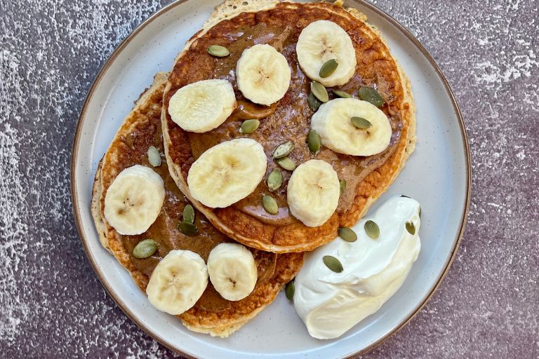 Pancakes on a plate with a spoon of Greek yoghurt on the side. Pancakes are toped with sliced banana and pumpkin seeds