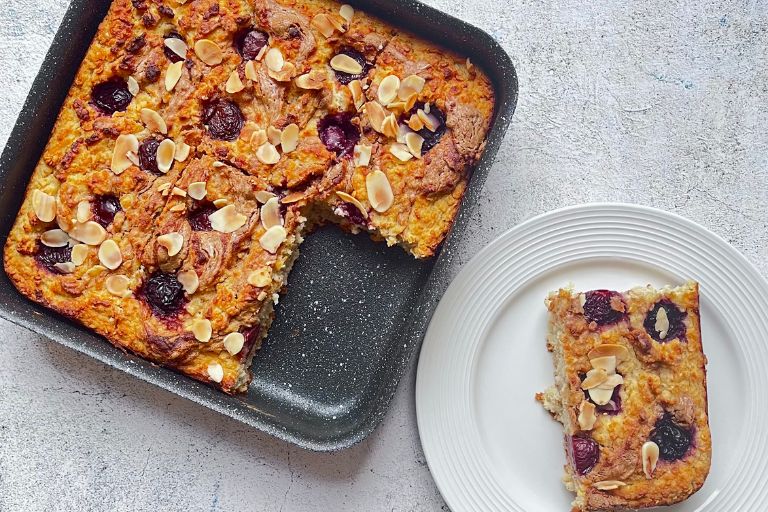 Oat, Pear and Cherry Breakfast Bake in a Baking Tray with a slice cut out which is on a white plate next to baking tray.