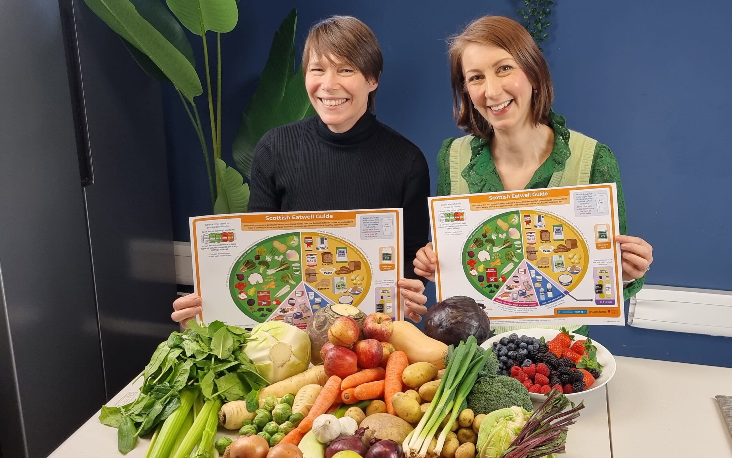 Suzanne and dr Laura Wyness are holding the first page of the Scottish Eatwell guide. Suzanne is wearing black sweater and dr Laura Wyness is in a green shirt. In front of them there is a table with Scottish fruits and vegetables including onions, potatoes, broccoli, blueberries, strawberries and more