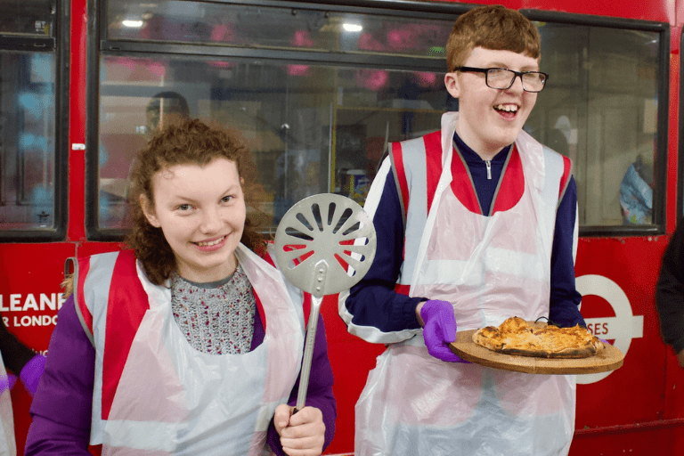 two teenagers standing in front of a red bus. They are wearing aprons,looking at the camera and smiling. A girl is holding a spatula and a boy is holding a plate with baked pie