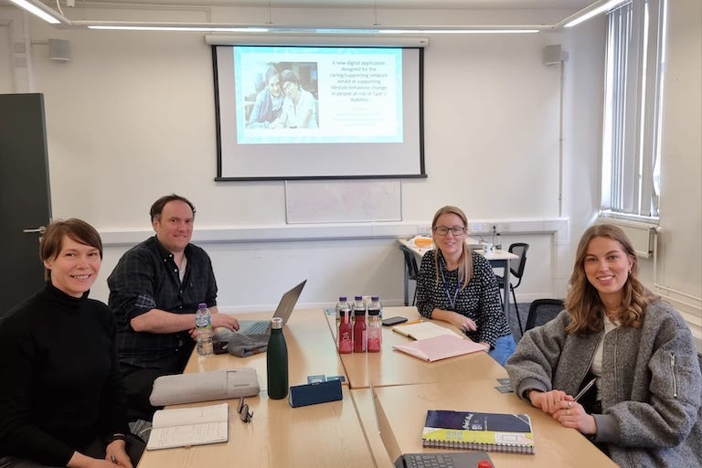 3 women and one man sitting at the desk with the laptop and notebooks, and projector screen behind them. They are smiling and looking at the camera