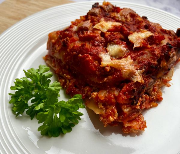 A serving of lasagna on a white plate, garnished with a sprig of fresh parsley with a rich tomato sauce and melted cheese on top