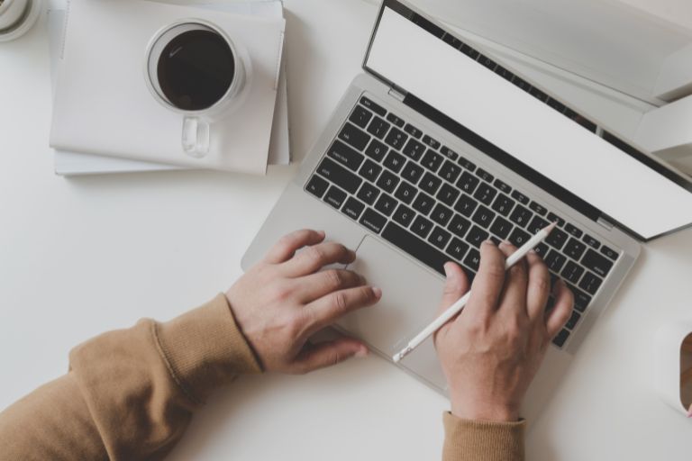 two hands using a laptop, the right hand is also holding a pen. There is a cup of black coffee to the side of the laptop